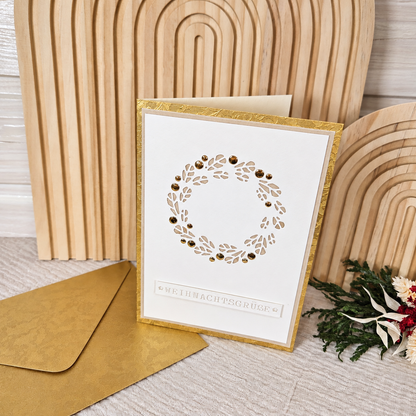 Weihnachtskarte „Weihnachtsgrüße“ mit goldfarbenem Umschlag vor einem hellen Holzhintergrund