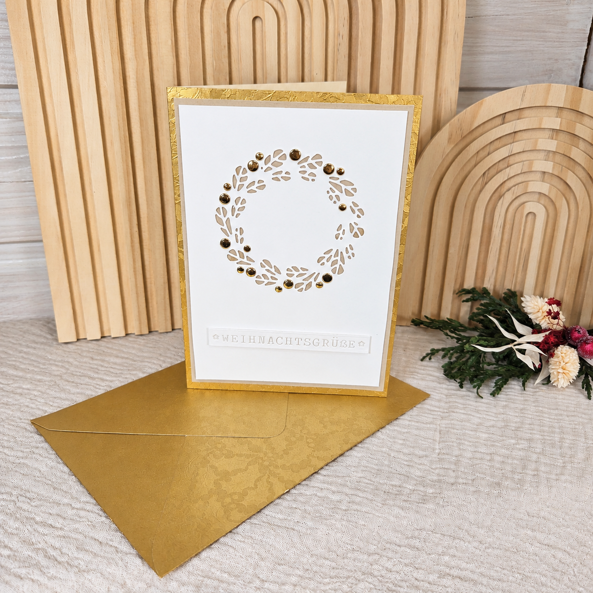 Aufrecht stehende Weihnachtskarte mit filigranem Kranz und goldenem Umschlag in weihnachtlicher Dekoration