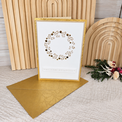 Aufrecht stehende Weihnachtskarte mit filigranem Kranz und goldenem Umschlag in weihnachtlicher Dekoration
